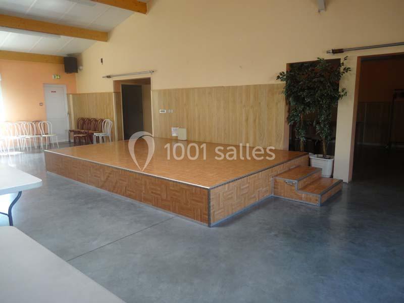 Petite estrade en bois dans une salle avec murs beige, sol en béton, chaises empilées et plante en pot.