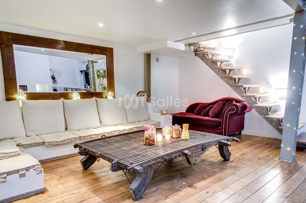 Salon lumineux avec canapé beige, table basse en bois, fauteuil rouge et escalier moderne en arrière-plan.