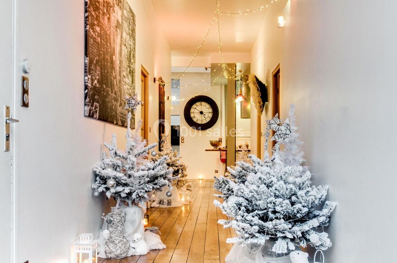 Couloir décoré avec des sapins enneigés, guirlandes lumineuses et lanternes, menant à une pièce éclairée.
