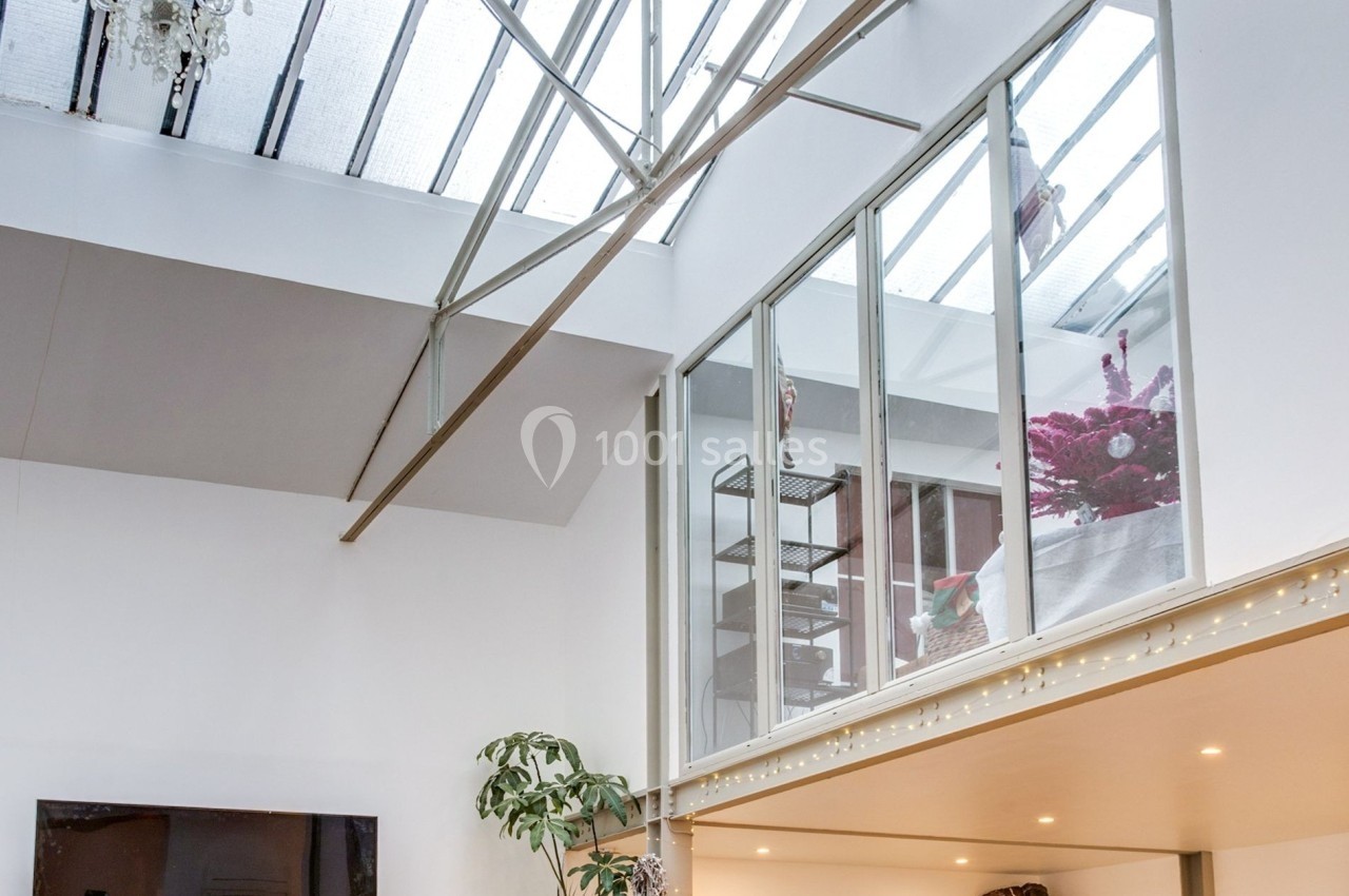 Salon lumineux avec parquet, grande verrière, canapé, plantes, télévision murale et mezzanine.