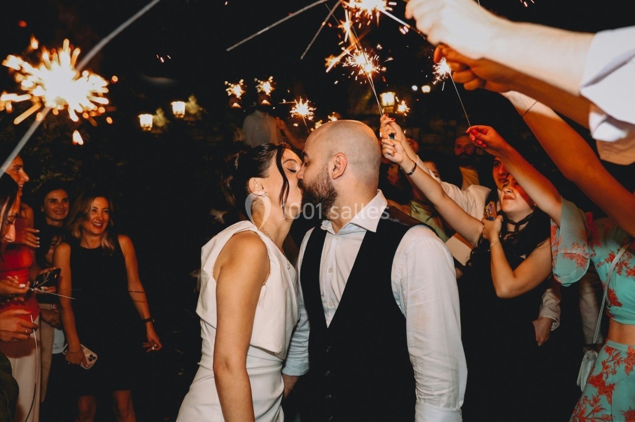 Un couple s'embrasse au centre d'une fête nocturne, entouré d'invités tenant des cierges magiques allumés.