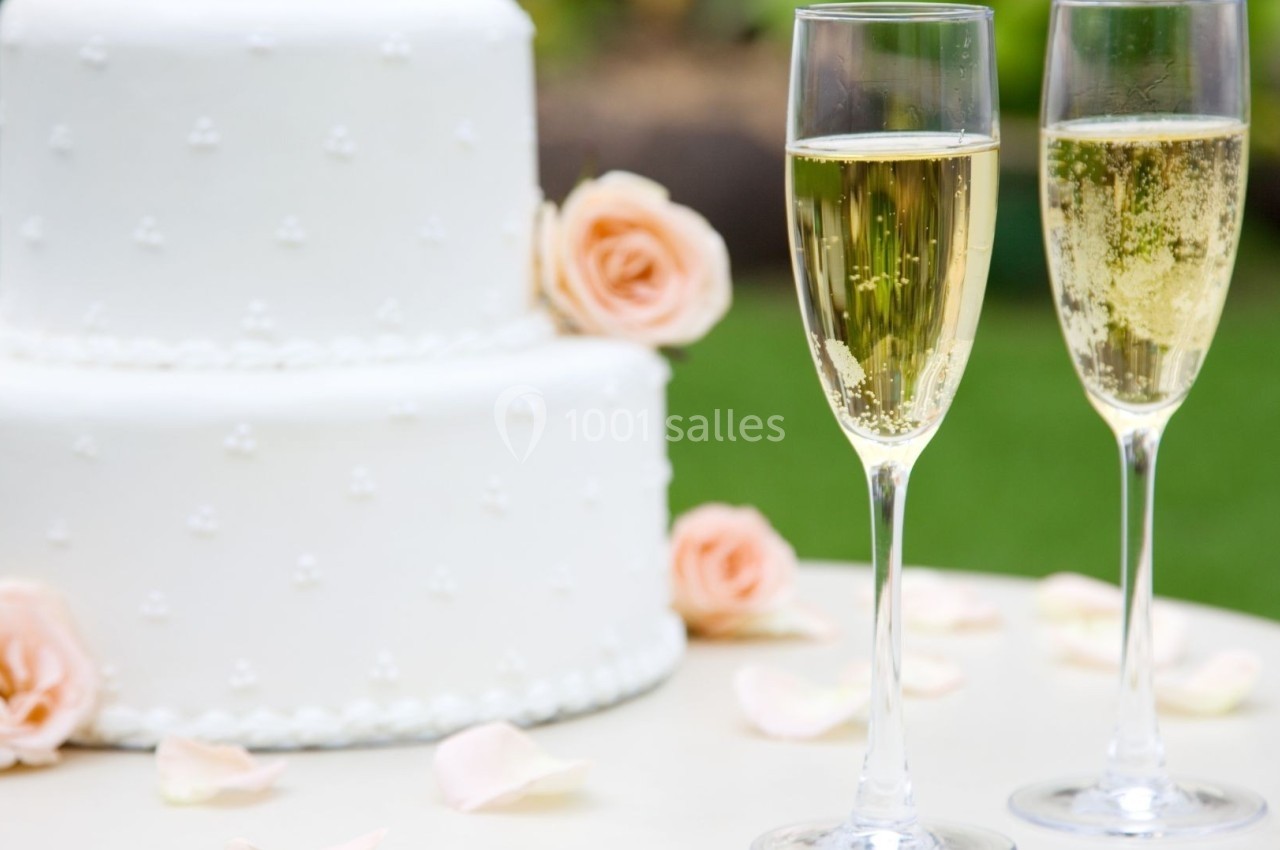 Gâteau de mariage blanc décoré de perles et deux coupes de champagne sur une table avec des pétales de rose.