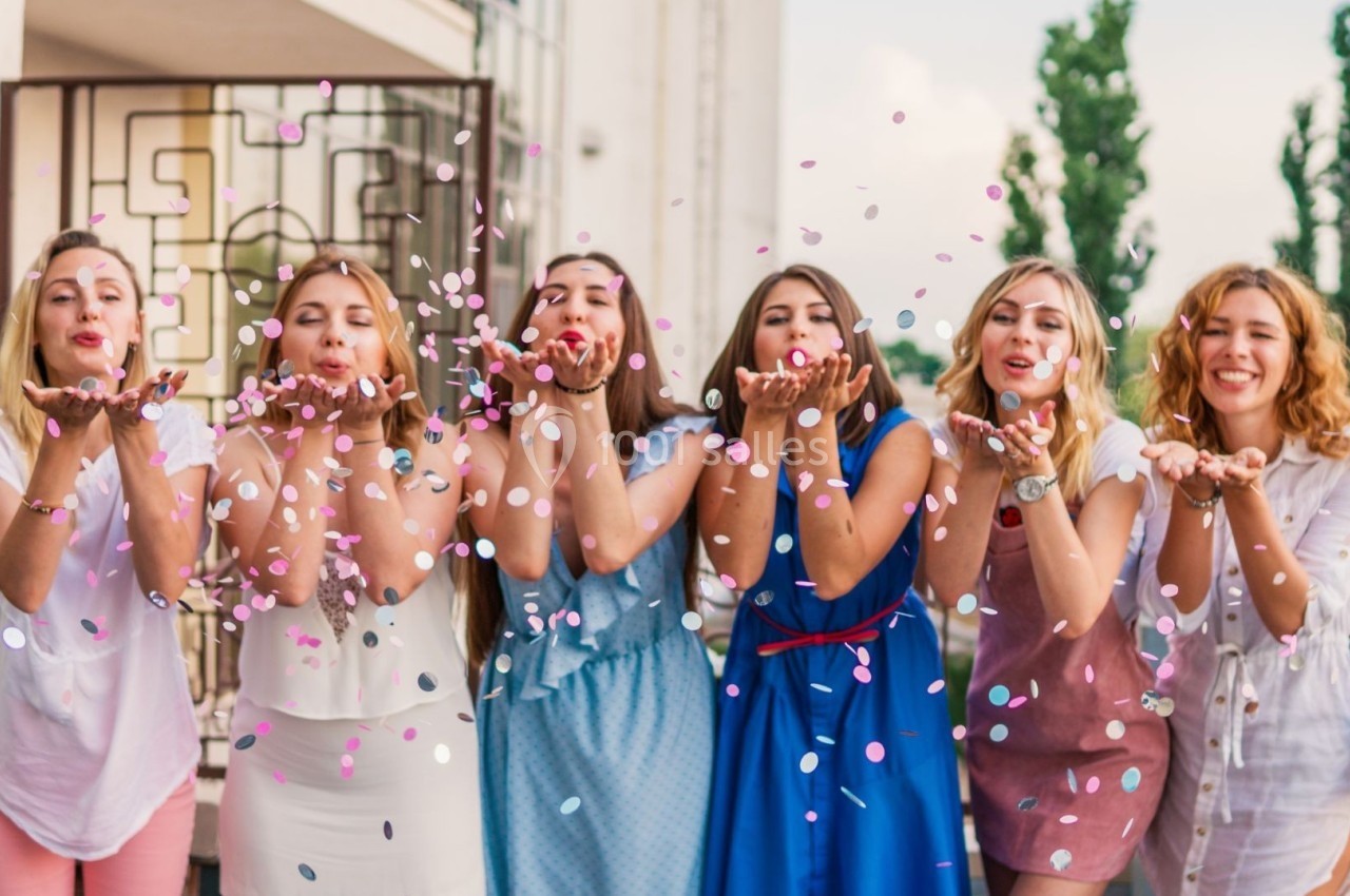 Six femmes souriantes debout sur une terrasse, soufflant des confettis colorés vers la caméra.
