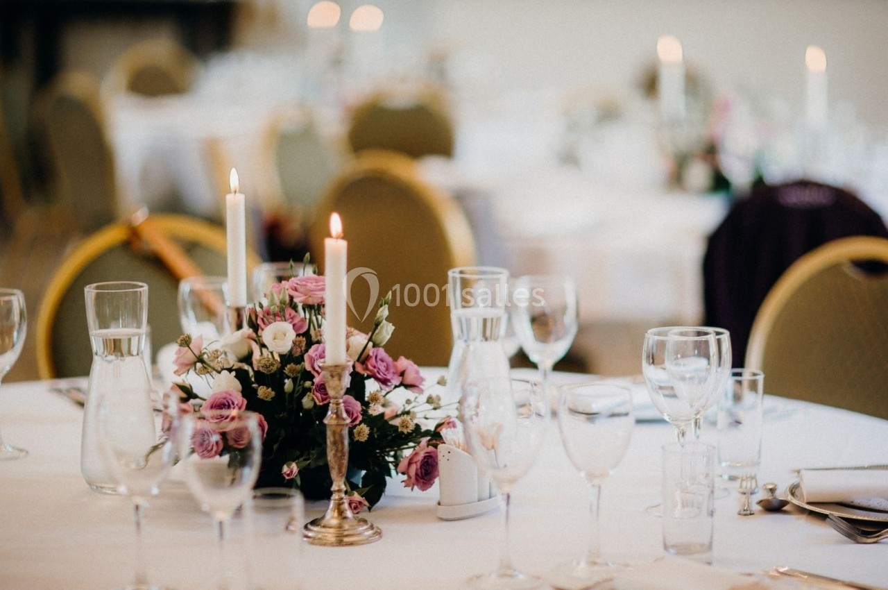 Table dressée avec des verres, des couverts, un centre de table floral et des bougies allumées dans une salle lumineuse.