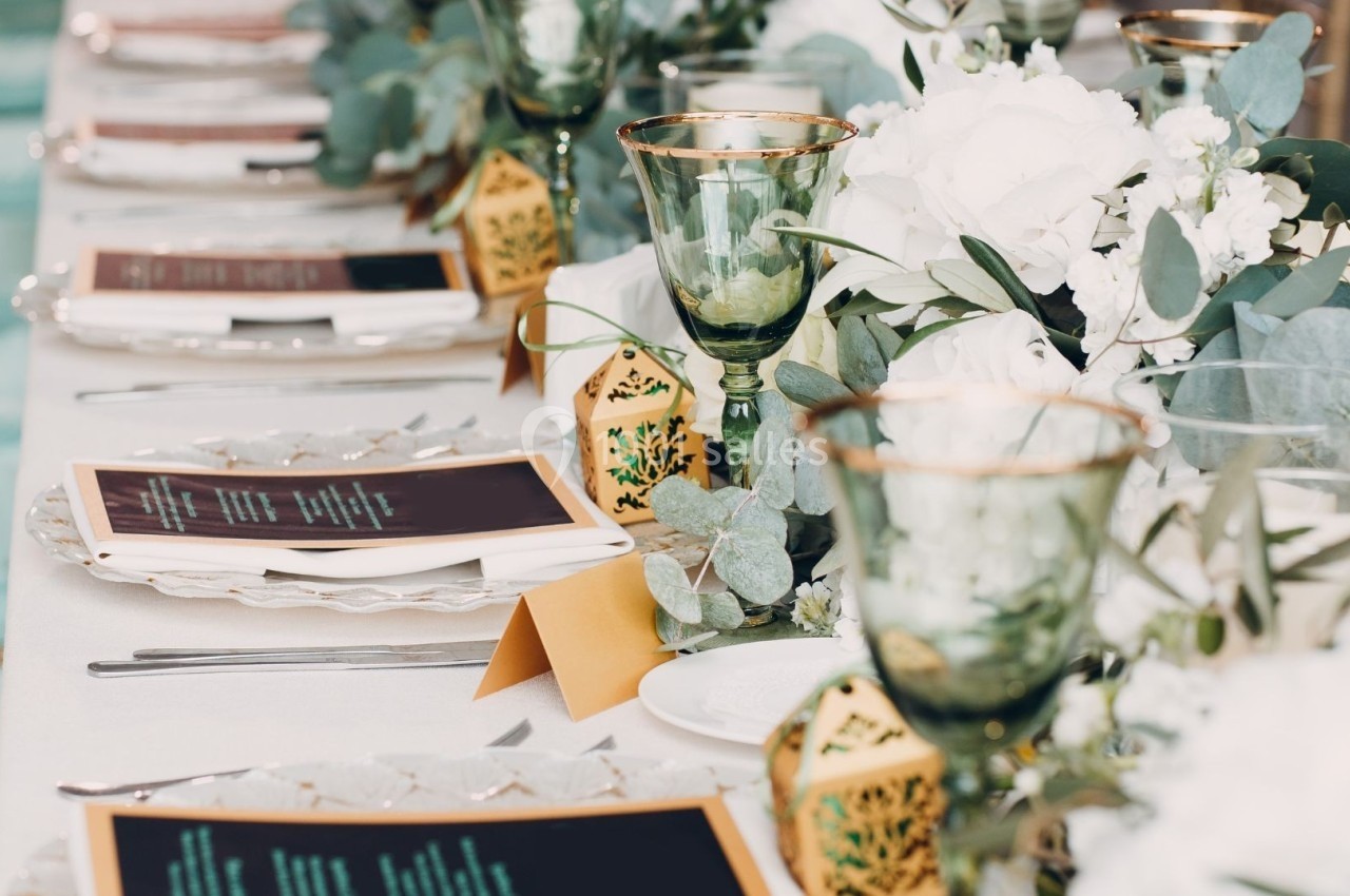 Table élégamment dressée avec des menus, des verres dorés, des fleurs blanches et des décorations végétales.