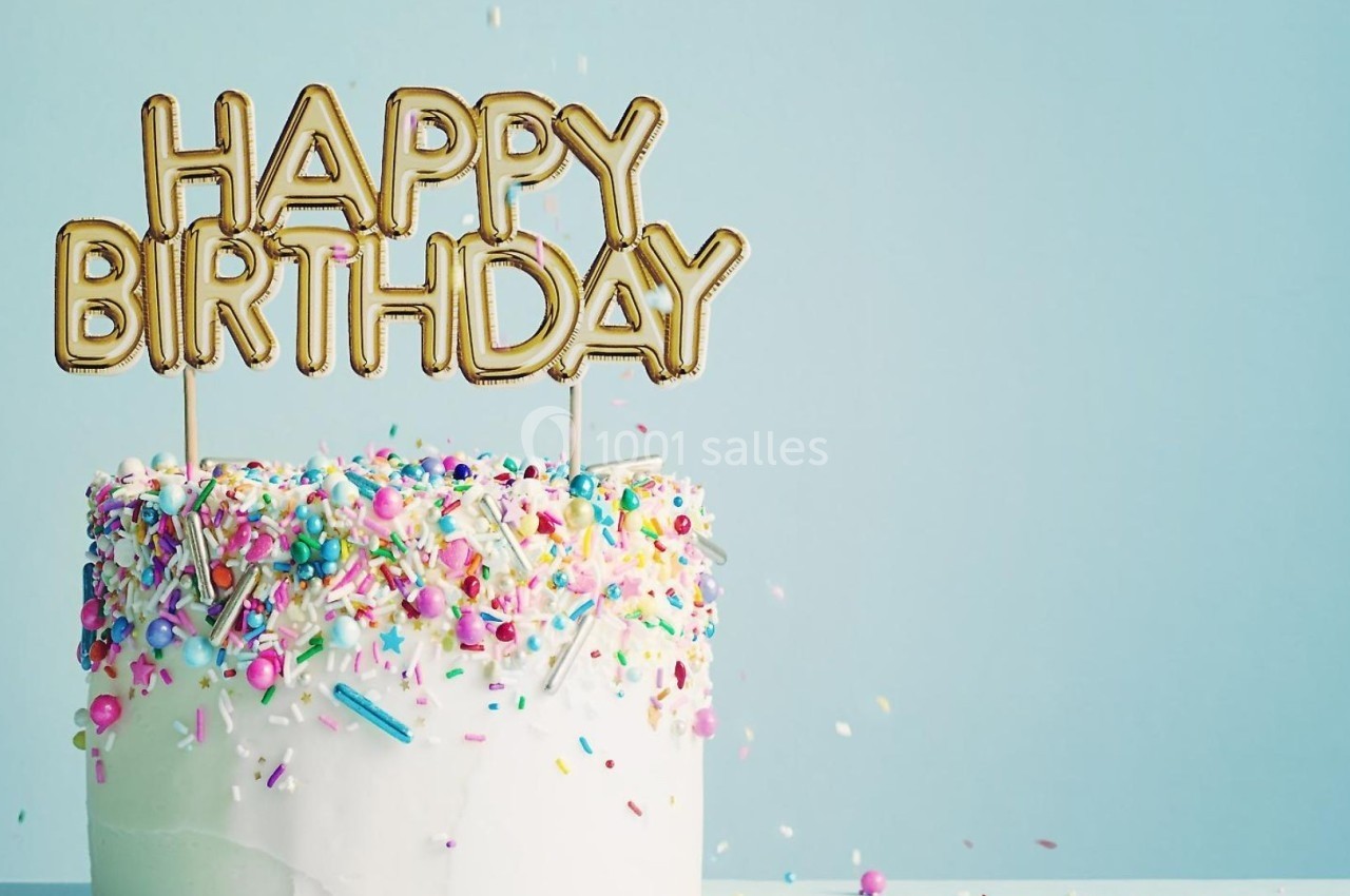 Gâteau d'anniversaire décoré de glaçage blanc, de confettis colorés et de ballons dorés formant ’Happy Birthday’.
