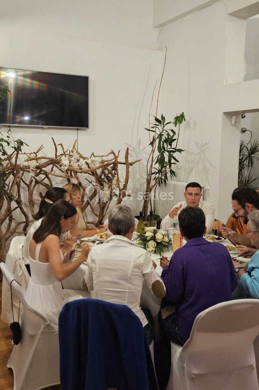 Des personnes assises autour d'une table décorée, partageant un repas dans une salle lumineuse avec des branches en arrière…