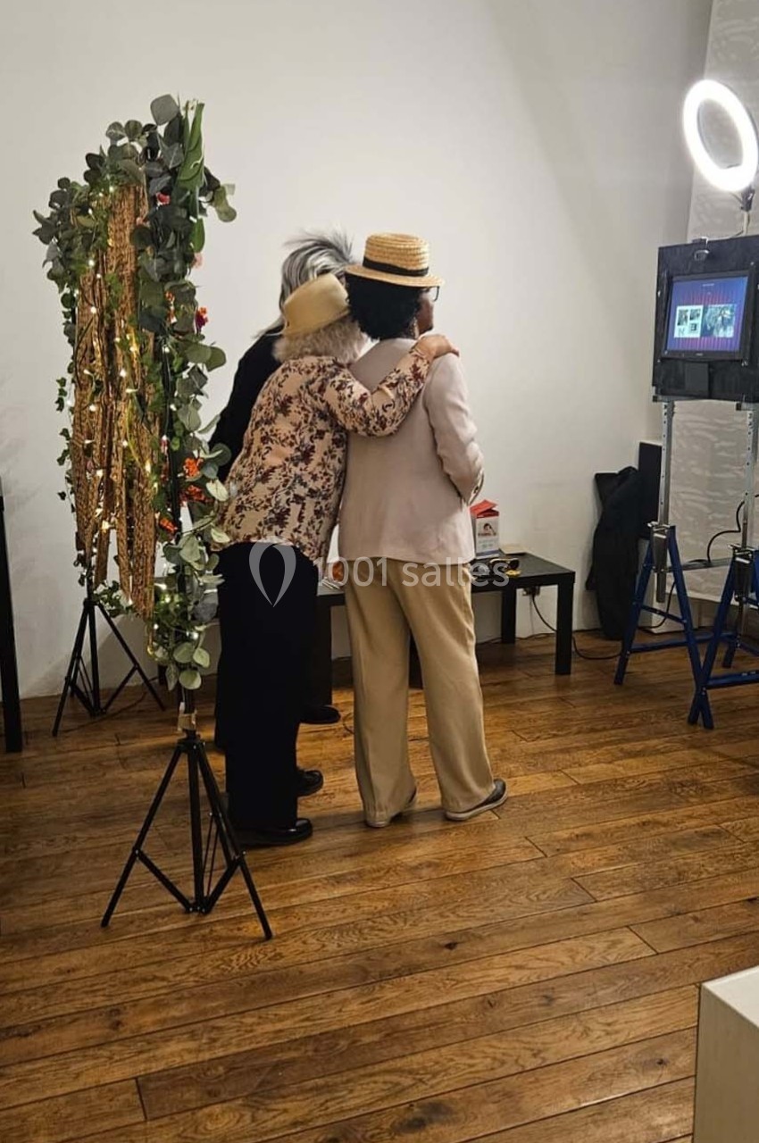 Deux personnes de dos, portant des chapeaux, posent devant un décor floral et un éclairage de studio.