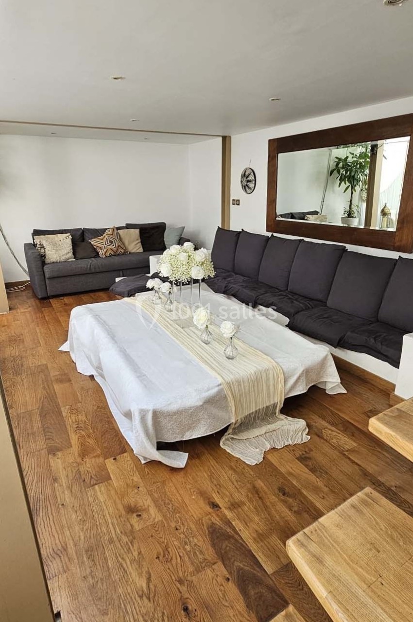 Salon avec parquet, table basse décorée de fleurs blanches, canapés gris et grand miroir mural.