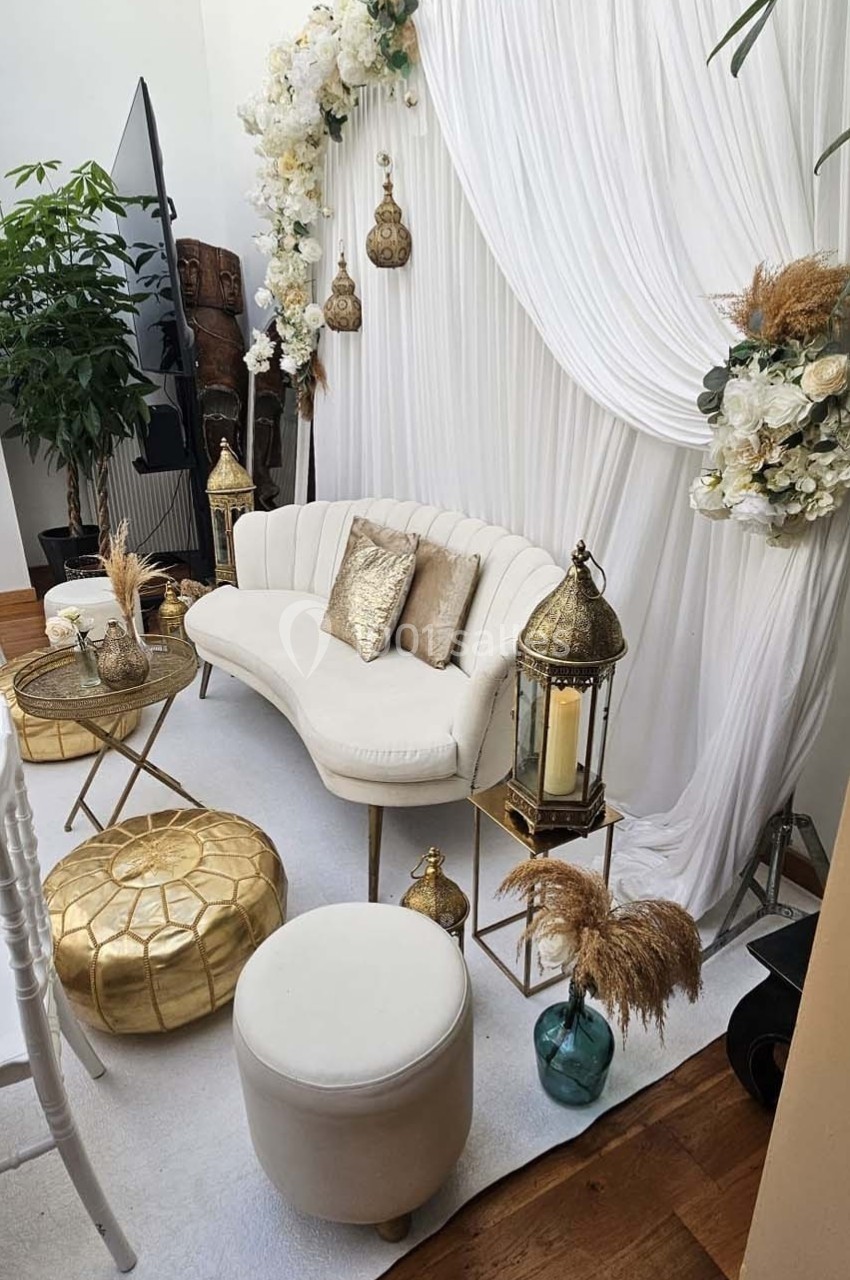 Salon décoré avec un canapé blanc, coussins dorés, lanternes, fleurs blanches et rideaux drapés.