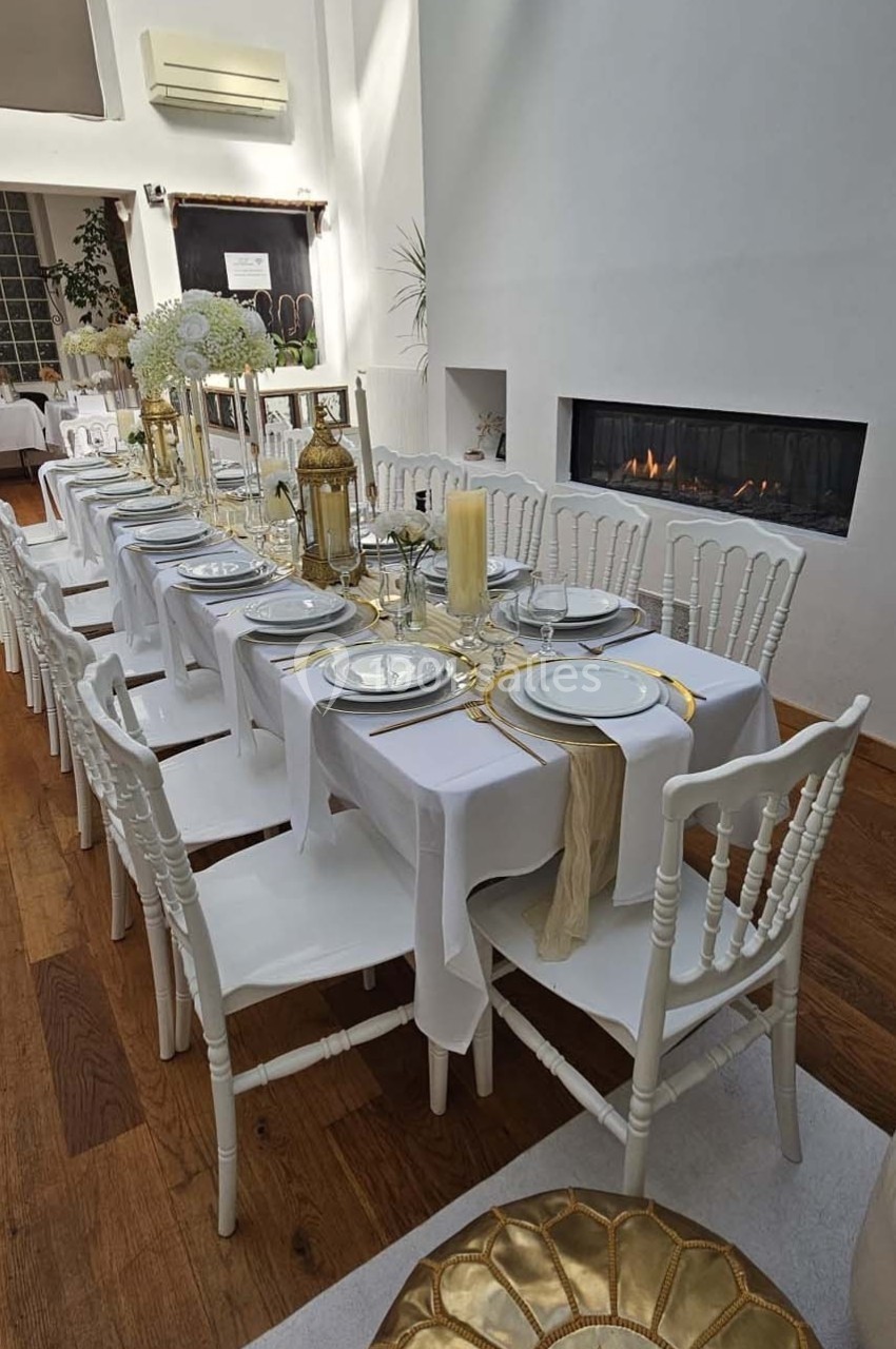 Table dressée avec nappes blanches, vaisselle élégante, bougies et décorations dorées dans une salle lumineuse avec cheminée.