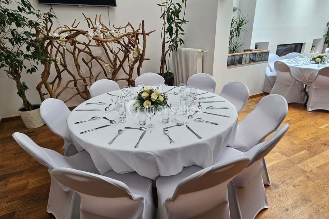 Salle décorée avec des tables rondes dressées, nappes blanches, chaises assorties et centre de table floral.