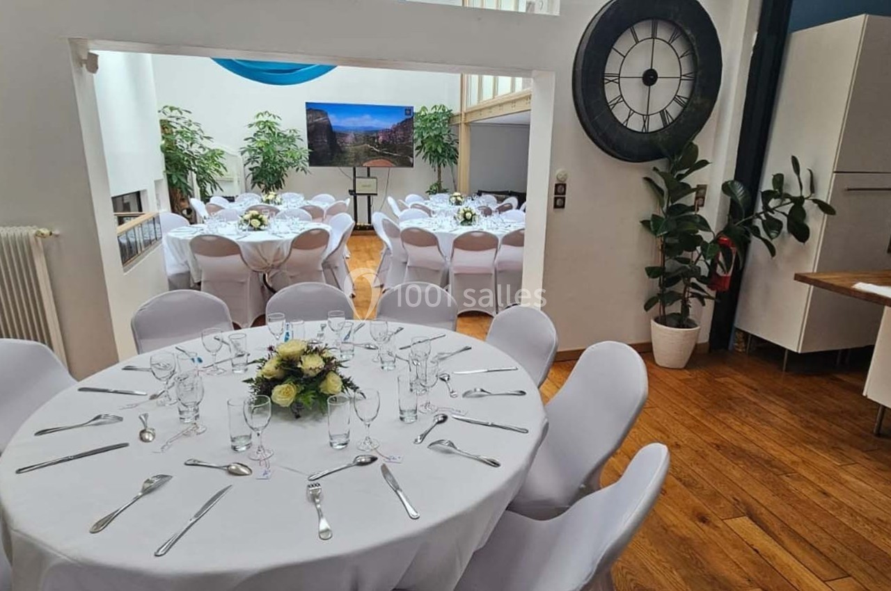 Salle lumineuse aménagée pour un événement, avec tables rondes dressées, plantes et grande horloge murale.