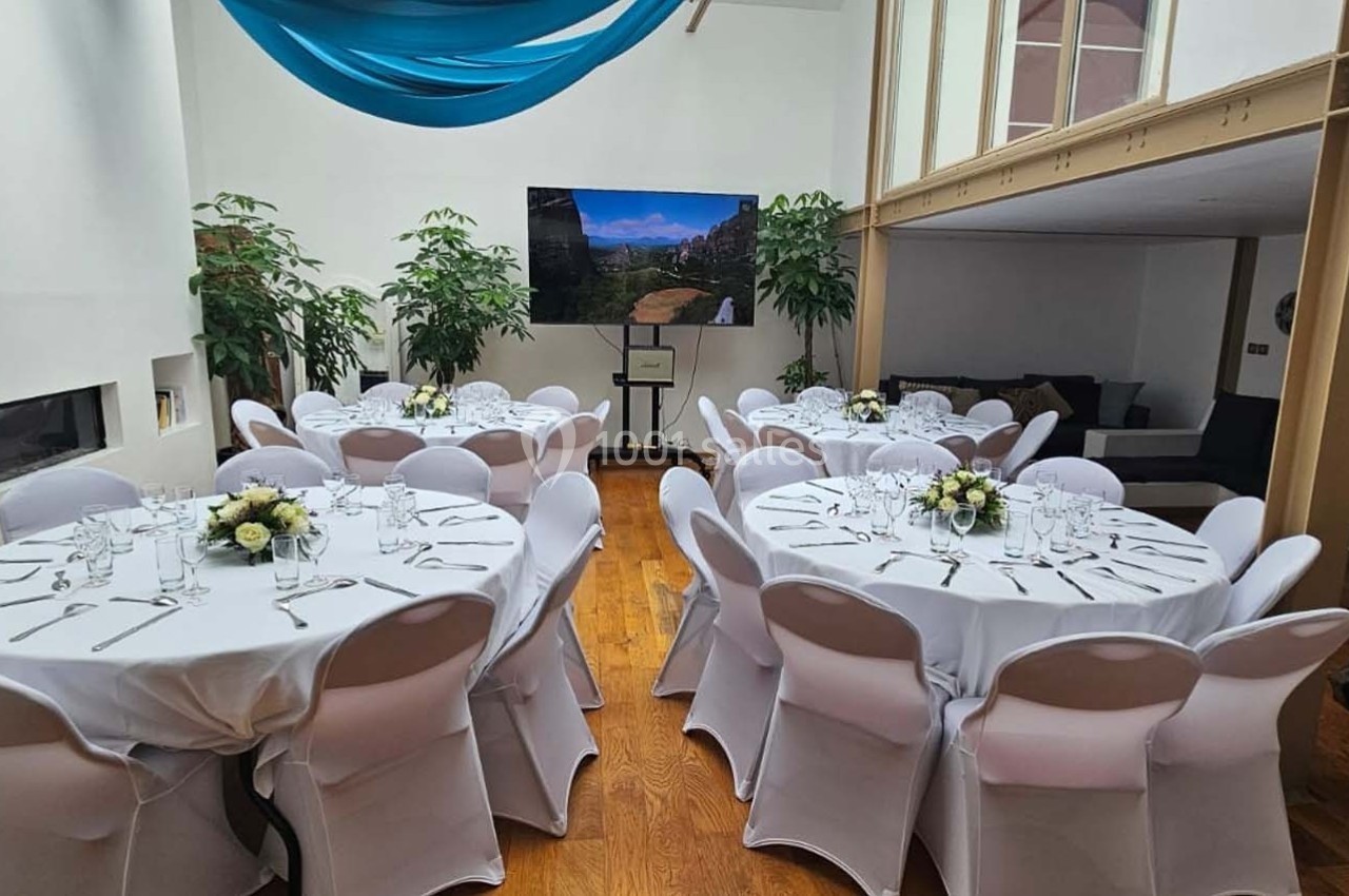 Salle lumineuse avec tables rondes dressées, nappes blanches, décor floral et écran affichant un paysage.