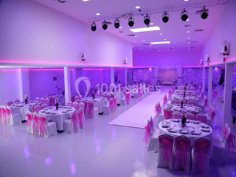 Salle de réception décorée avec des tables rondes, nappes blanches et chaises ornées de rubans roses, éclairage violet.