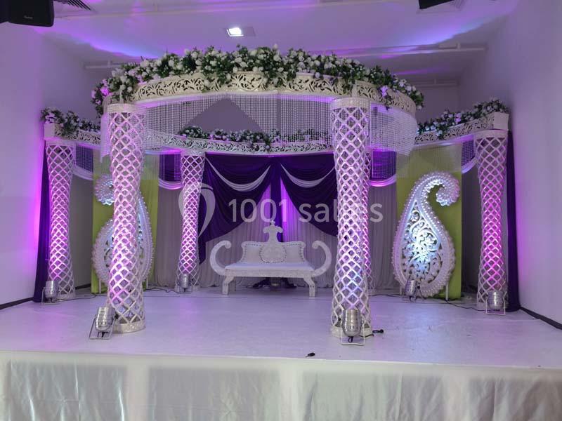 Décor de scène de mariage avec arche ornée de fleurs blanches, rideaux violets et canapé blanc sculpté.