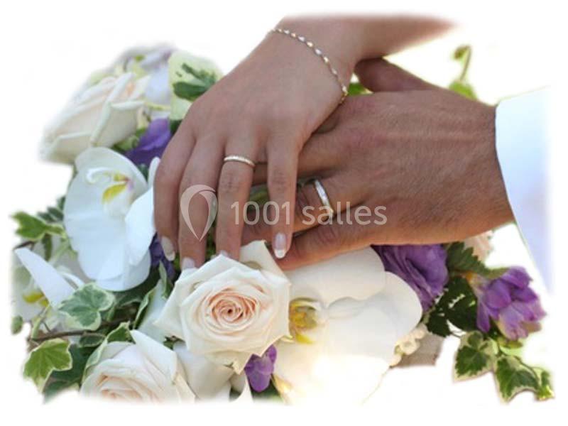 Mains d'un couple portant des alliances, posées sur un bouquet de fleurs blanches et violettes.