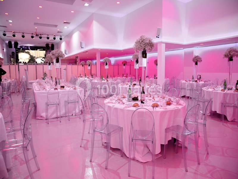 Salle de réception décorée avec des tables rondes, nappes blanches, chaises transparentes et éclairage rose.