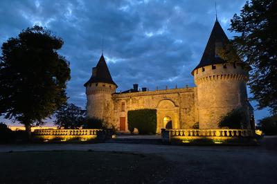 Château en pierre éclairé à la tombée de la nuit, entouré d'arbres et surmonté d'un ciel nuageux.