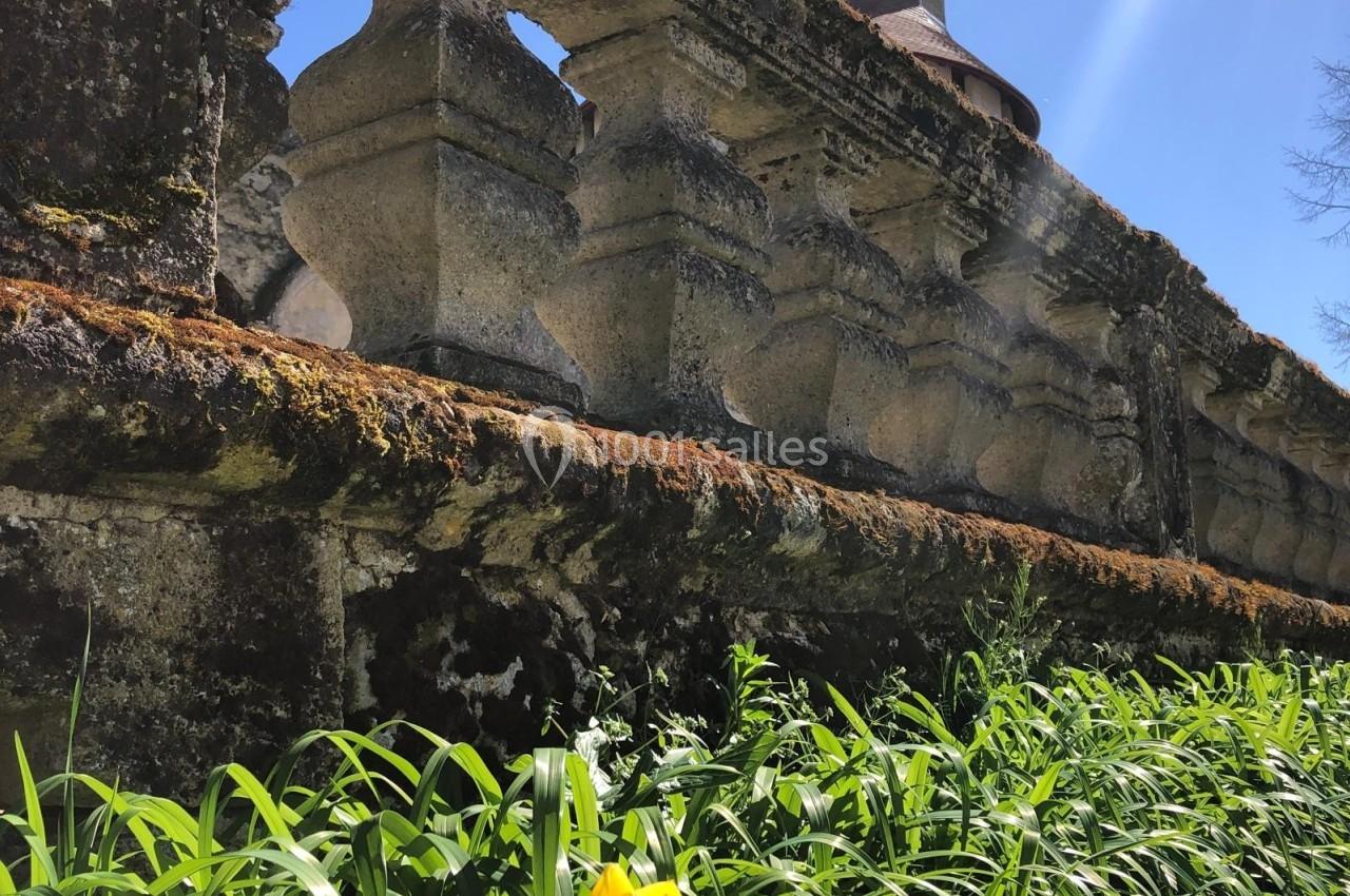 Mur en pierre ancienne avec mousse, surmonté de balustres, entouré de verdure et d'une tulipe jaune au premier plan.