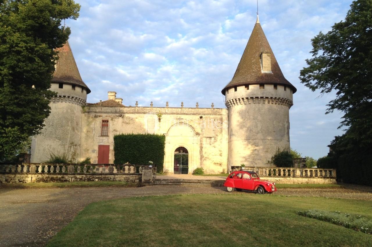 Château ancien avec deux tours rondes, pelouse au premier plan et une petite voiture rouge stationnée devant.