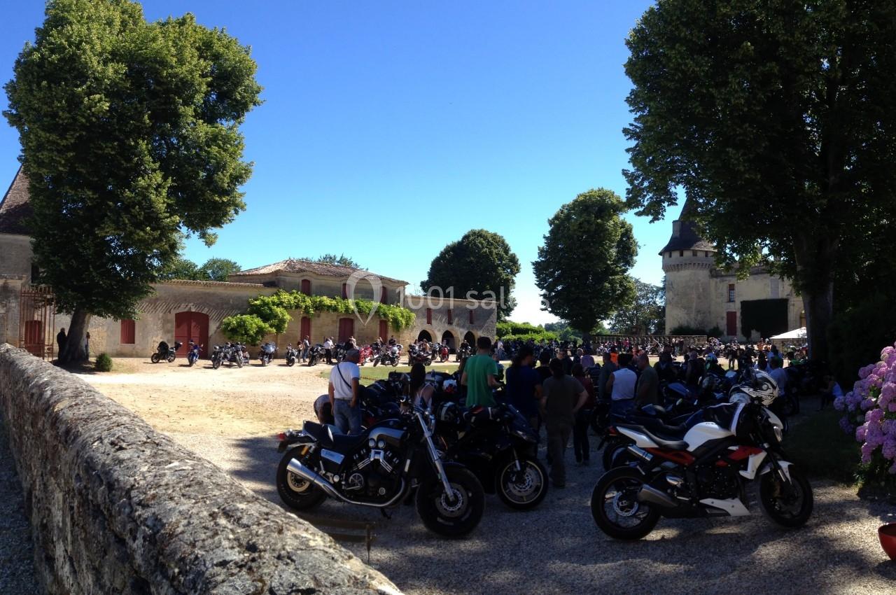 Rassemblement de motos et de participants dans la cour d'un domaine entouré d'arbres par une journée ensoleillée.
