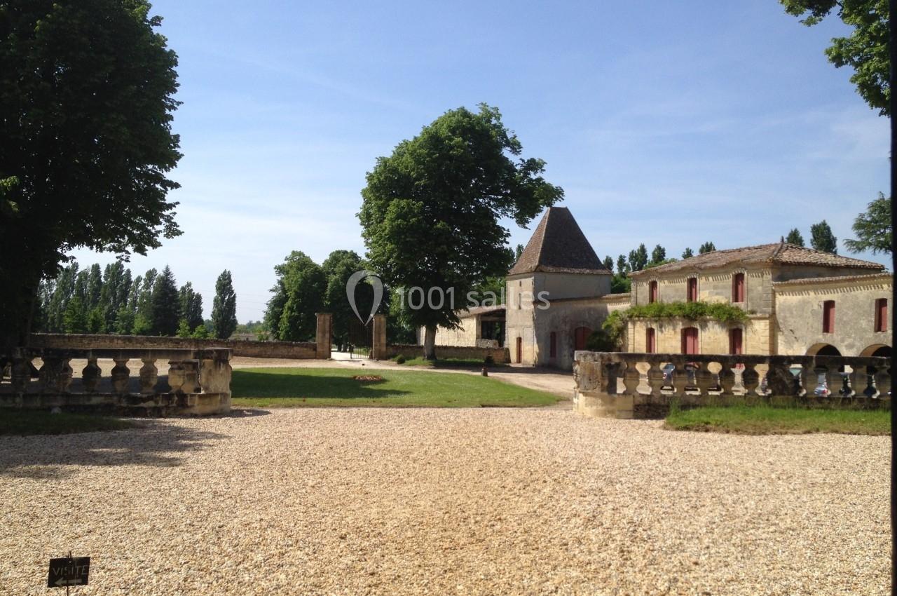 Cour d’un domaine avec bâtiments en pierre, allée gravillonnée, balustrades et arbres sous un ciel dégagé.