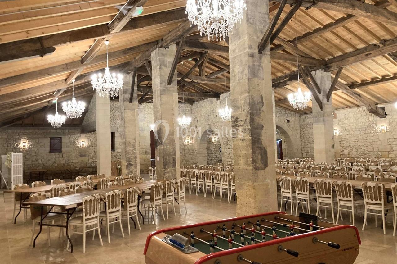 Salle en pierre avec poutres apparentes, lustres suspendus, tables et chaises alignées, et un baby-foot au premier plan.