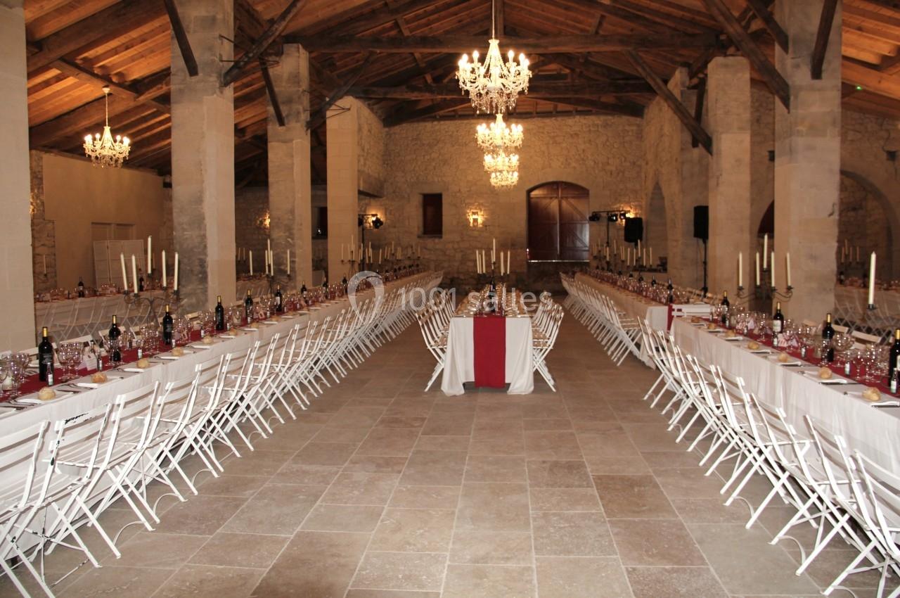 Grande salle de réception en pierre avec tables longues dressées, chaises blanches et chandeliers suspendus.
