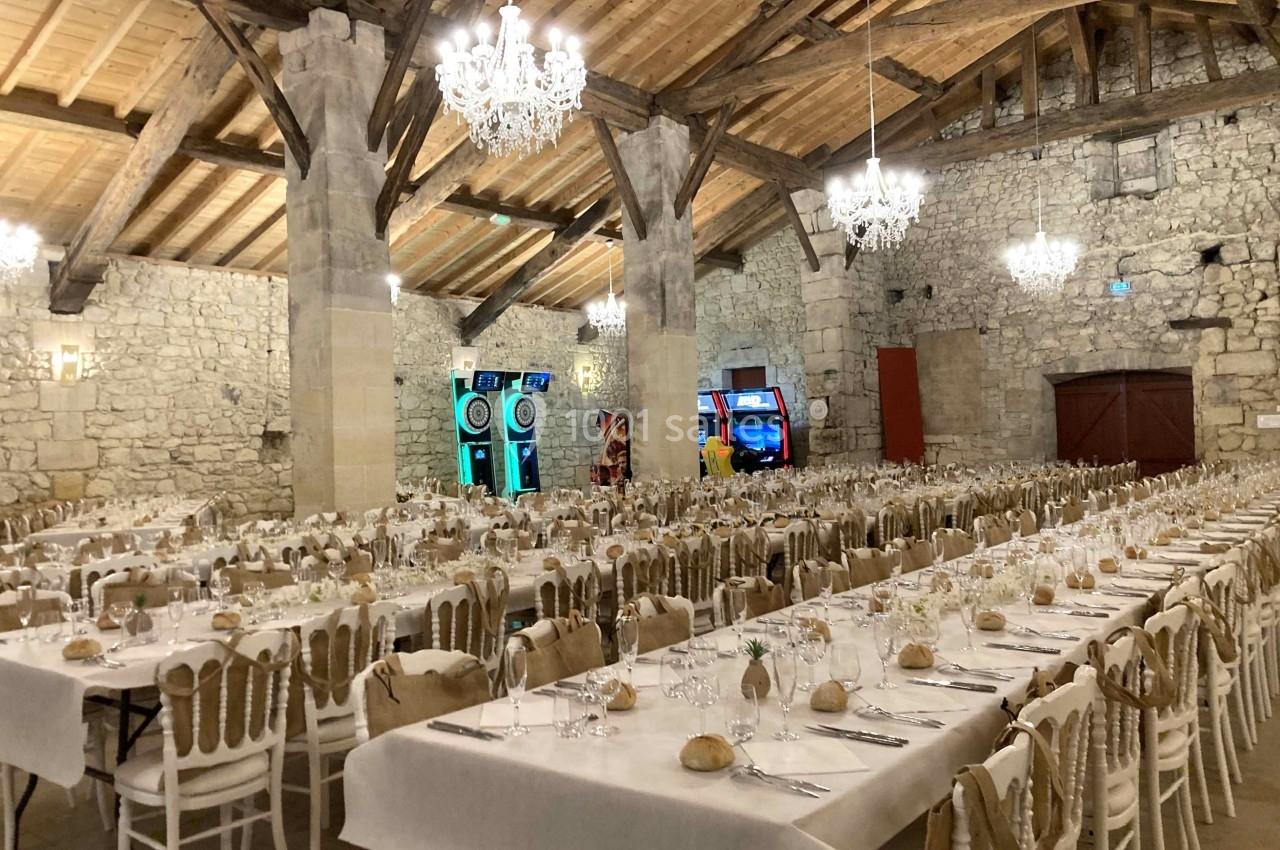 Salle de réception en pierre avec tables dressées, chaises en bois, lustres suspendus et machines de jeux au fond.
