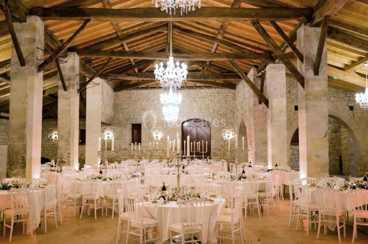 Salle de réception en pierre avec poutres apparentes, tables rondes dressées et lustres suspendus.