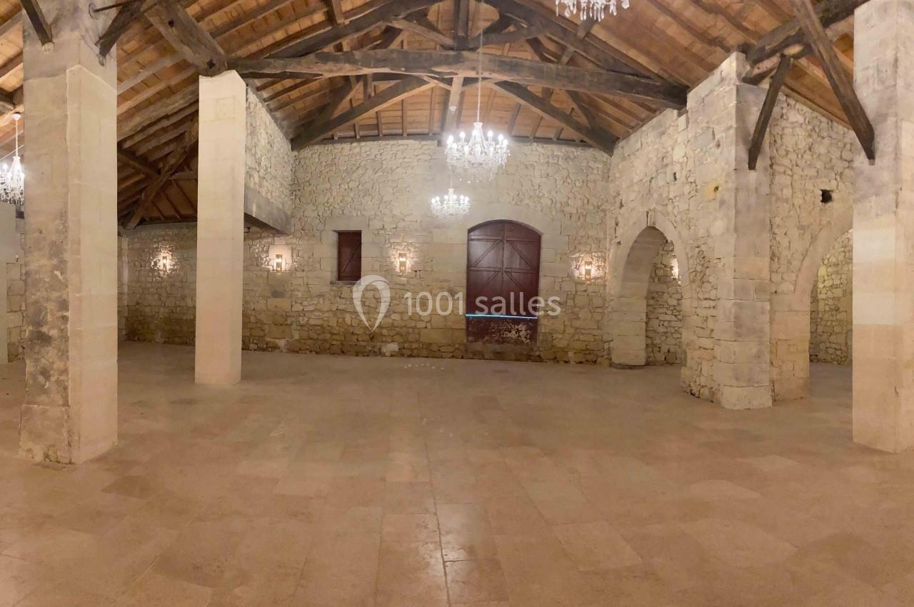 Salle en pierre avec poutres apparentes, sol en pierre claire, lustres suspendus et grandes ouvertures en bois.