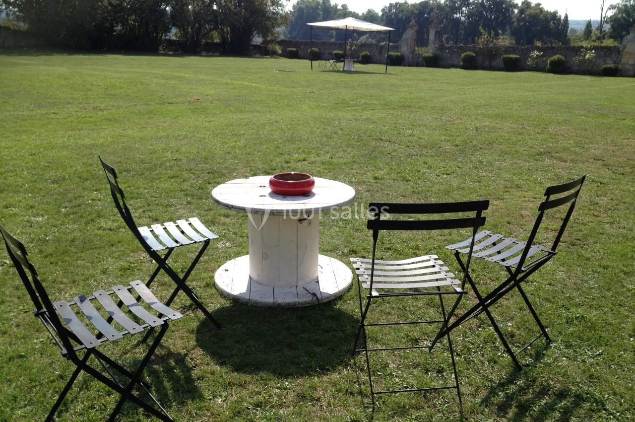 Quatre chaises noires entourent une table basse ronde en bois sur une pelouse, avec un parasol en arrière-plan.