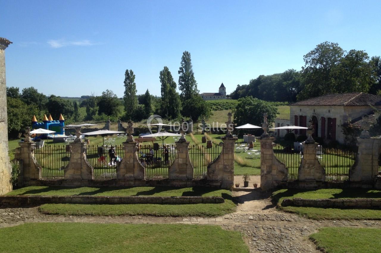 Vue d'un jardin avec pelouse, tables, parasols et une structure gonflable, entouré de bâtiments anciens et d'arbres.
