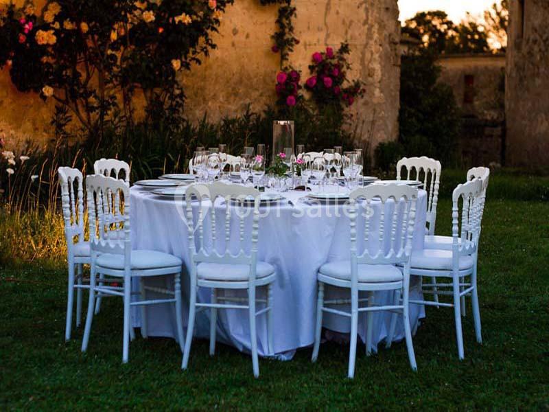 Table ronde dressée avec nappes blanches, chaises blanches et verres, installée dans un jardin au coucher du soleil.