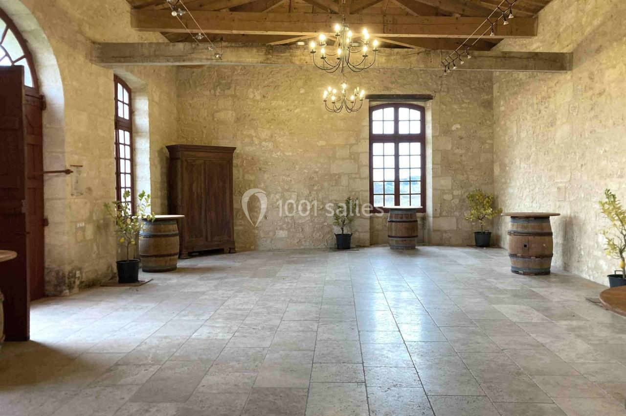 Salle en pierre avec plafond en poutres apparentes, fenêtres lumineuses, tonneaux en bois et plantes en pots.