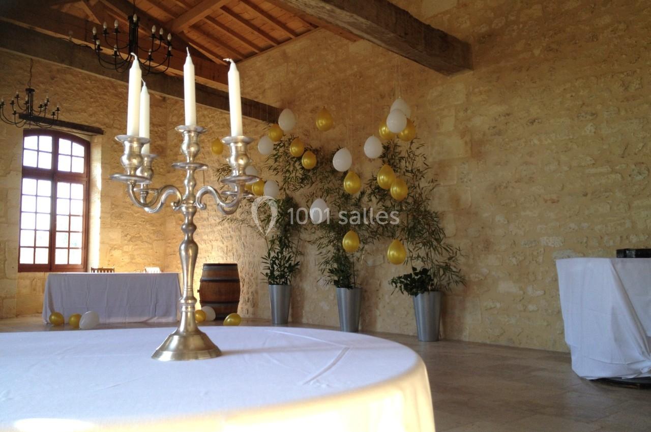 Salle en pierre avec plafond en bois, table décorée d'un chandelier, ballons jaunes et blancs accrochés au mur.