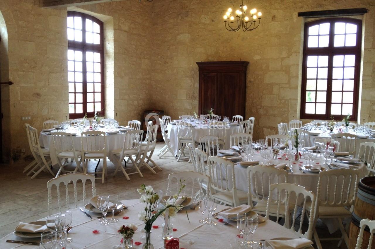 Salle de réception lumineuse avec tables rondes dressées, chaises blanches et décorations florales.