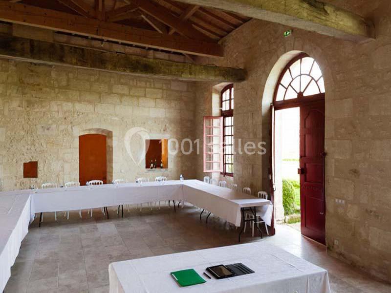Salle en pierre avec poutres apparentes, tables disposées en U, chaises blanches et lumière naturelle entrant par des…