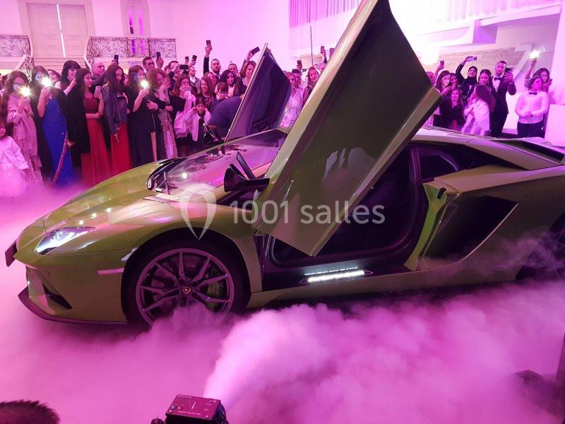 Voiture de sport verte aux portes papillon ouvertes, entourée de fumée et d'une foule dans un lieu éclairé.