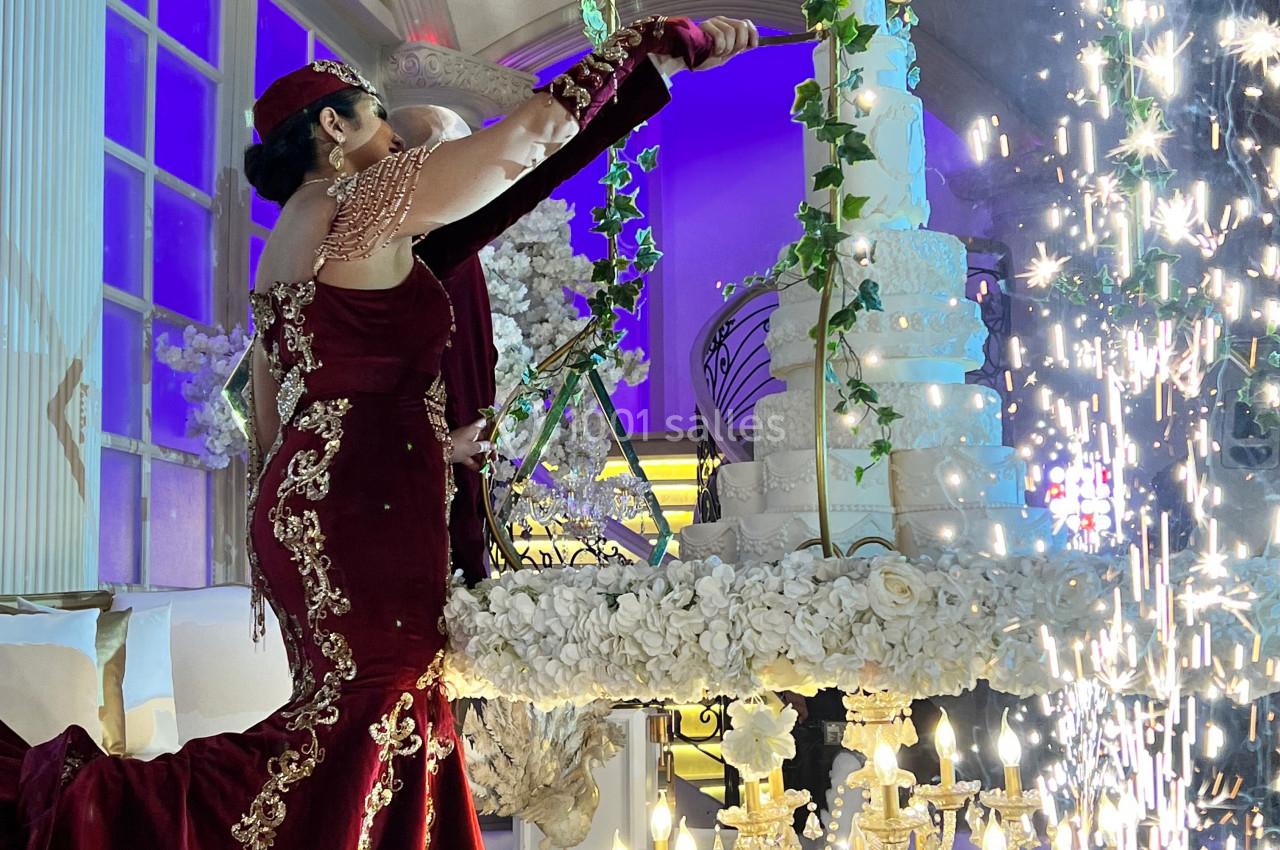 Une femme en robe rouge ornée de dorures ajuste un grand gâteau décoré de fleurs, entourée de chandeliers et d'étincelles.