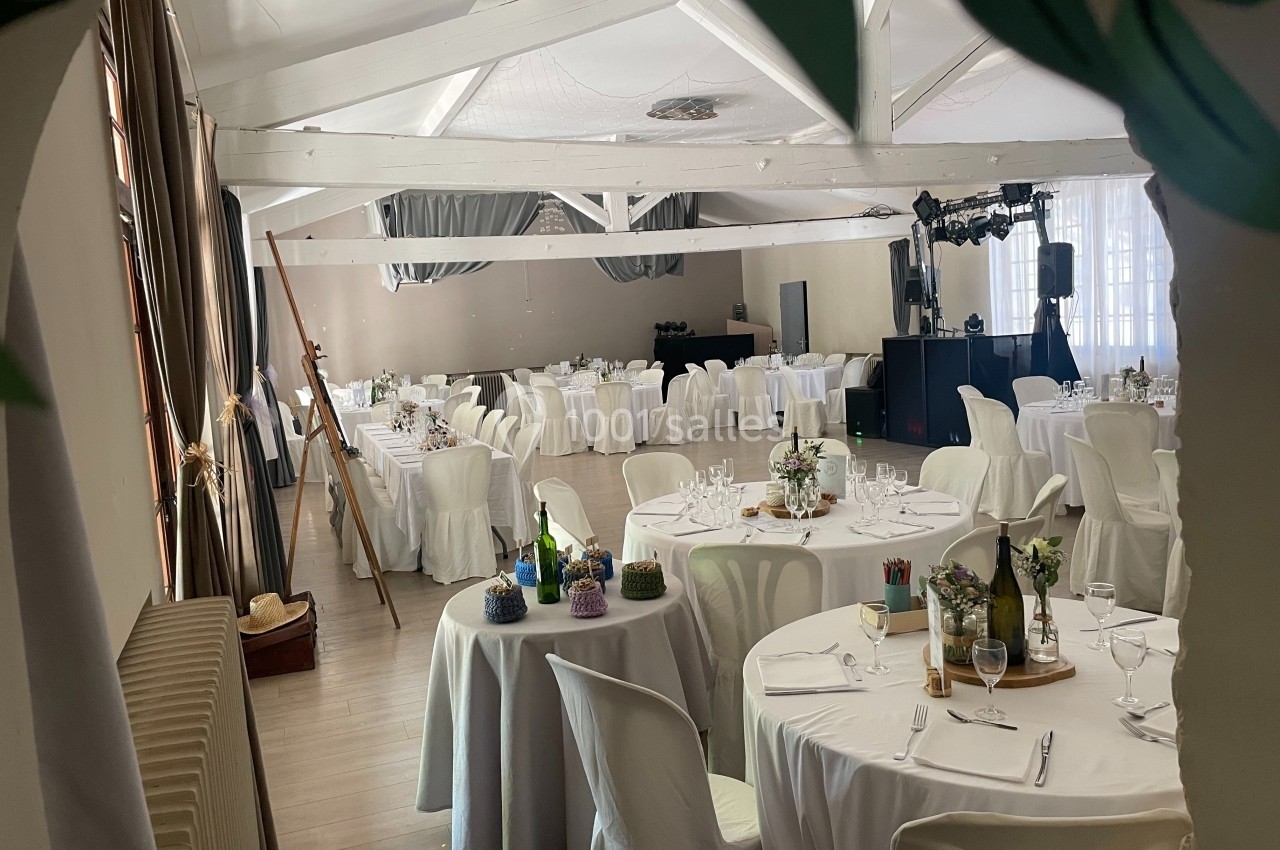 Salle de réception lumineuse avec tables rondes dressées, décorations florales et équipement de sonorisation.