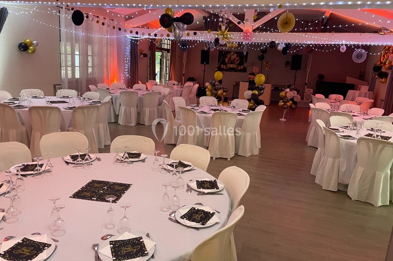 Salle décorée pour un événement festif avec tables rondes dressées, guirlandes lumineuses et ballons noirs et dorés.