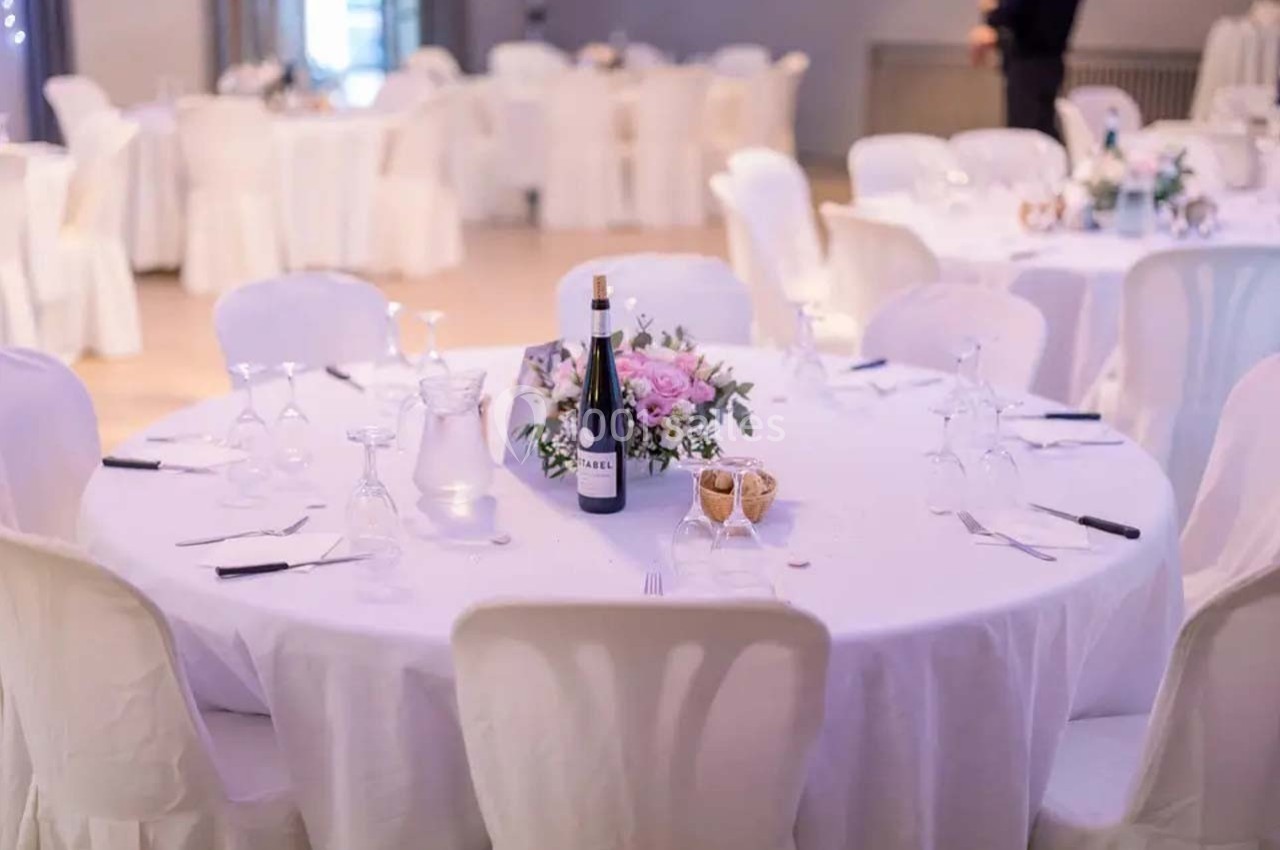 Table ronde dressée avec une nappe blanche, décorée de fleurs, verres, couverts et une bouteille de vin.