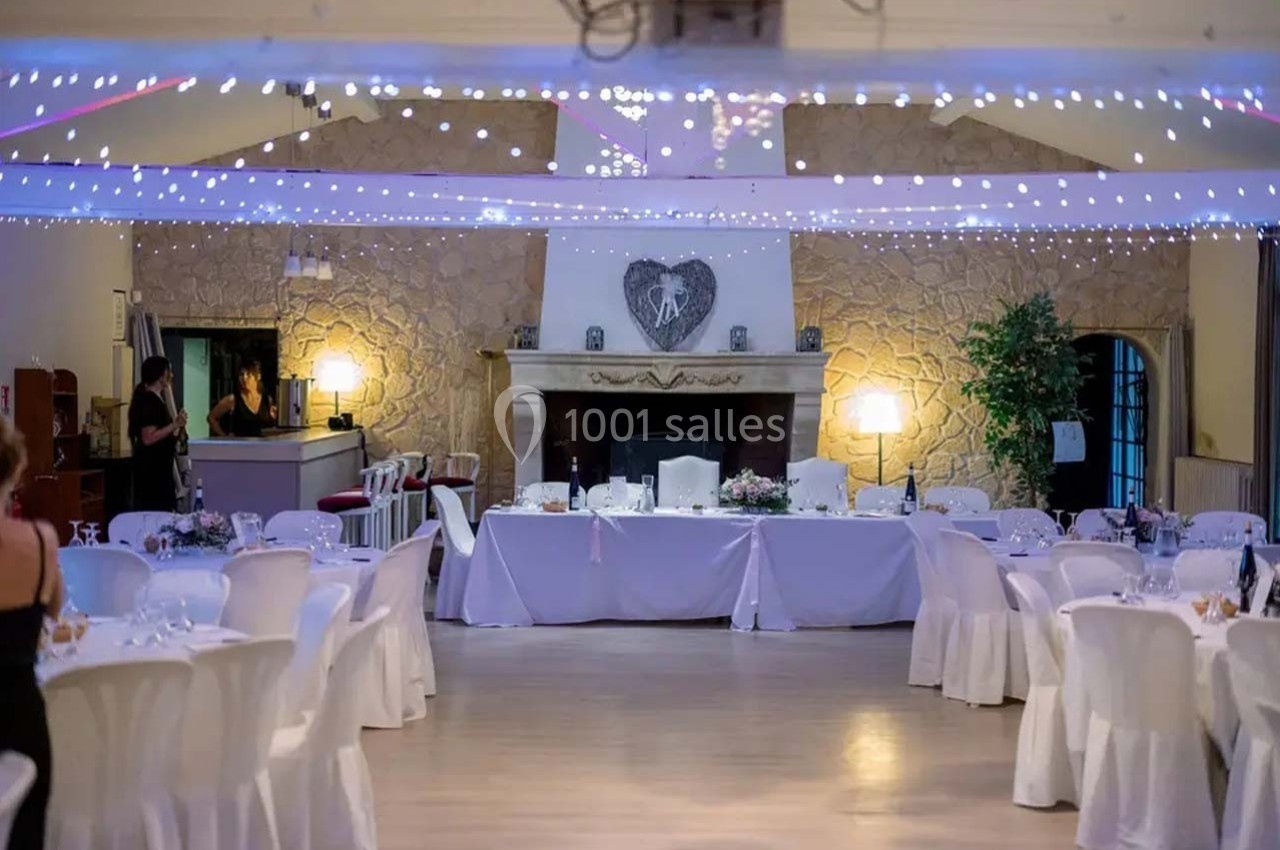 Salle de réception décorée avec des tables rondes, nappes blanches, guirlandes lumineuses et une cheminée en pierre.