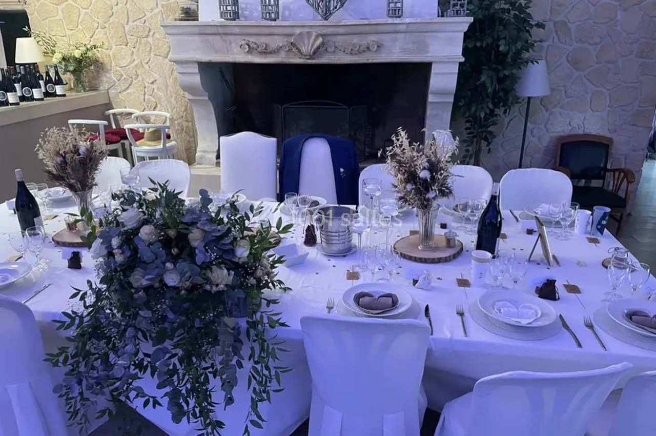 Table élégamment dressée pour un repas, décorée de fleurs séchées et entourée de chaises blanches, devant une cheminée en…
