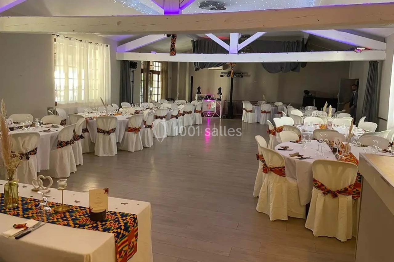 Salle de réception décorée avec des tables rondes, nappes blanches et chaises ornées de rubans colorés.