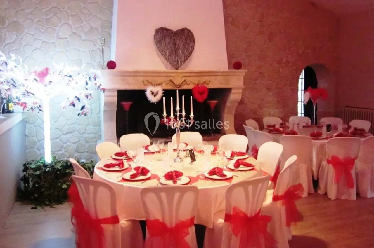 Salle décorée pour un événement, avec tables rondes dressées en blanc et rouge, et cheminée ornée d'un cœur en bois.