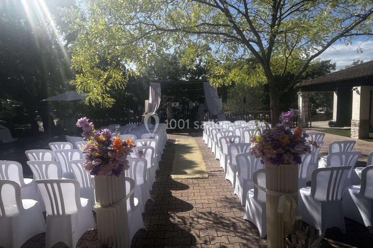 Chaises blanches alignées en extérieur, allée centrale décorée de fleurs menant à une arche sous un arbre.
