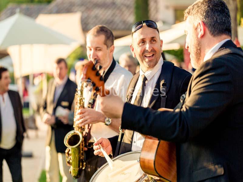 Des musiciens jouent du saxophone, de la guitare et du tambour en extérieur lors d'un événement ensoleillé.